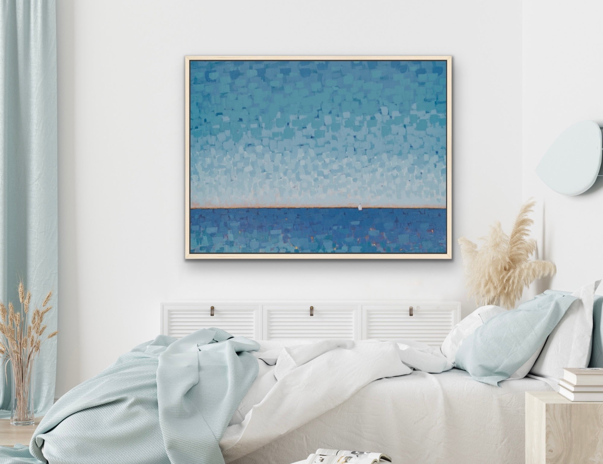 A framed square wall art print depicting a textured blue seascape hangs above a bed in a bright, modern bedroom with white bedding, light blue accents, and dried pampas grass.