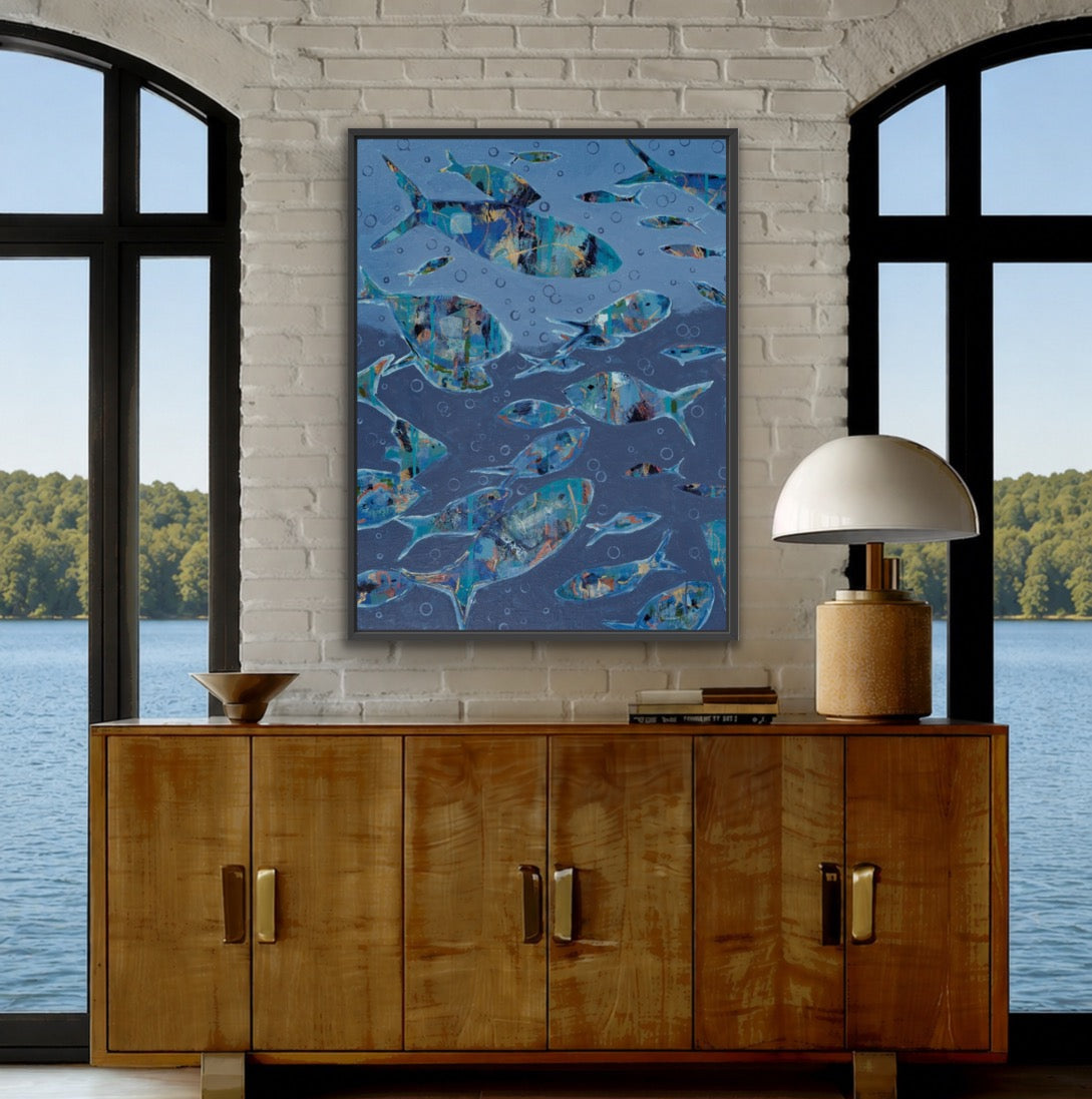 A vertical framed art print depicting a school of blue fish, displayed on a white brick wall above a wooden credenza in a modern, sunlit room with large windows overlooking water.