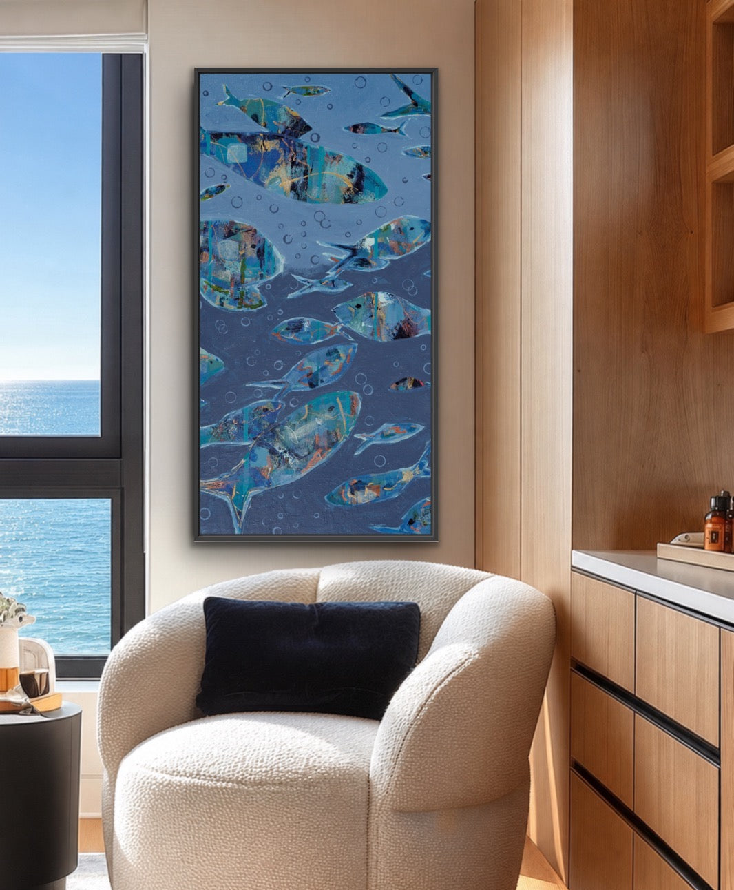 A framed vertical wall art print featuring a school of blue fish on a dark blue background, styled in a modern living room setting above a cream boucle armchair.