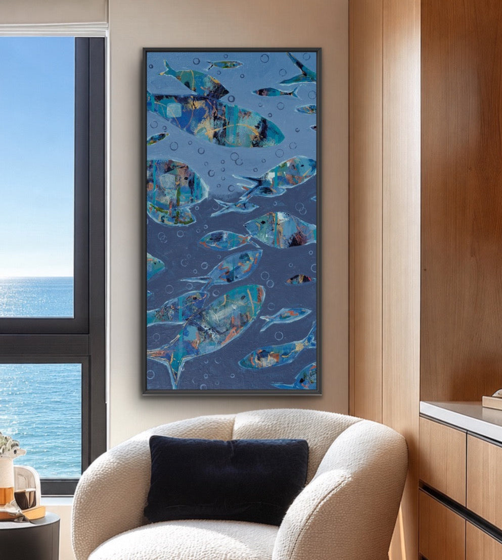 A framed vertical wall art print featuring a school of blue fish on a dark blue background, styled in a modern living room setting above a cream boucle armchair.