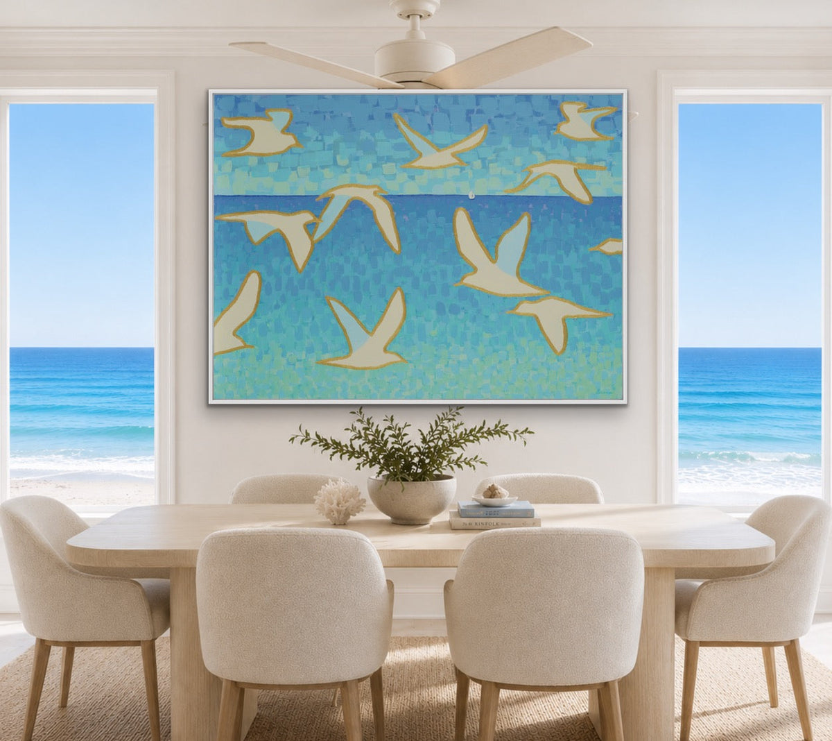 A large framed wall art print depicting flying seagulls in shades of blue and teal with cream accents, displayed in a bright, modern dining room with a table and chairs overlooking a beach and ocean view.