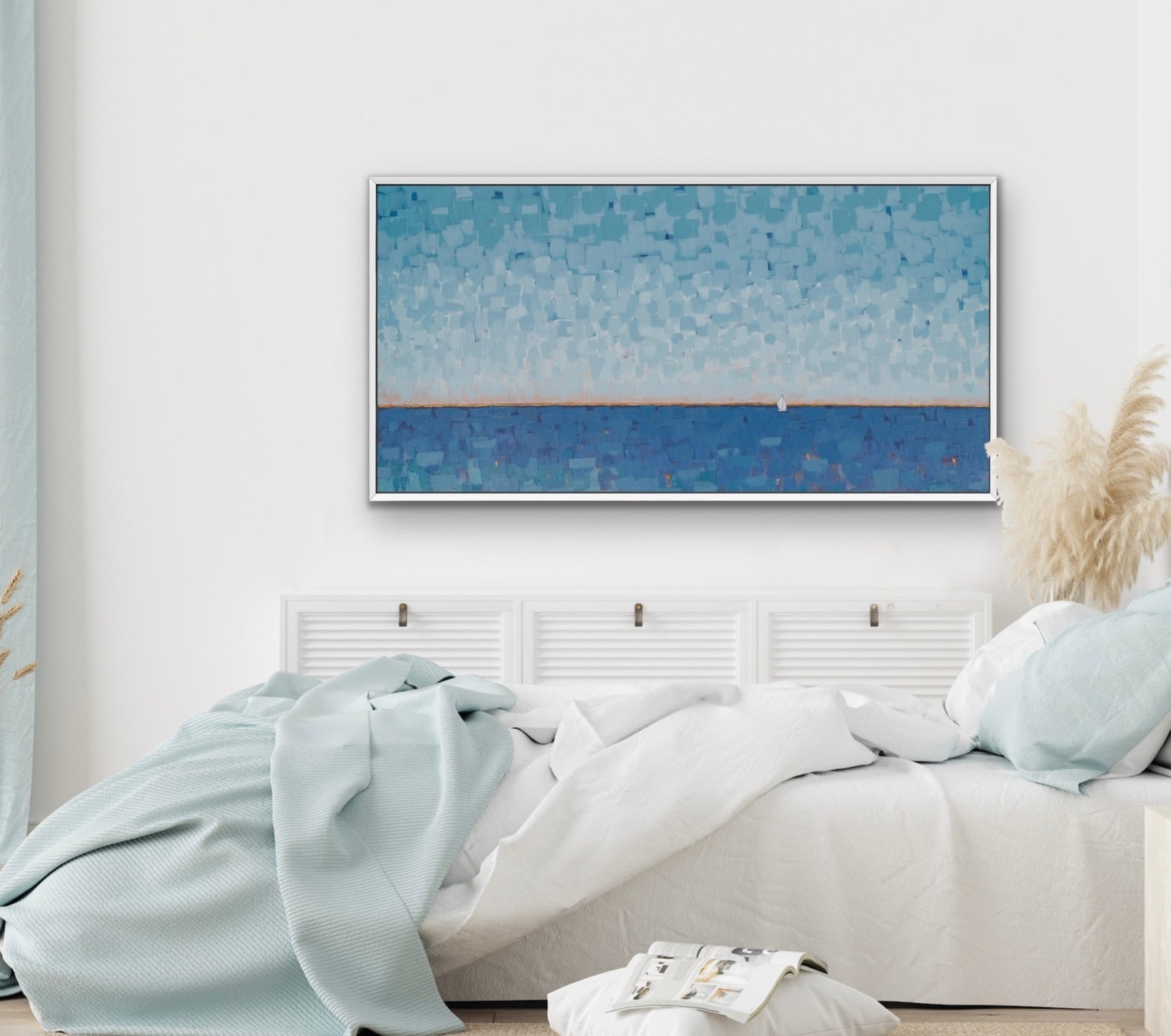 A framed horizontal canvas print of a textured blue seascape hangs on a white wall above a neatly made bed with white and light blue bedding in a bright, modern bedroom.