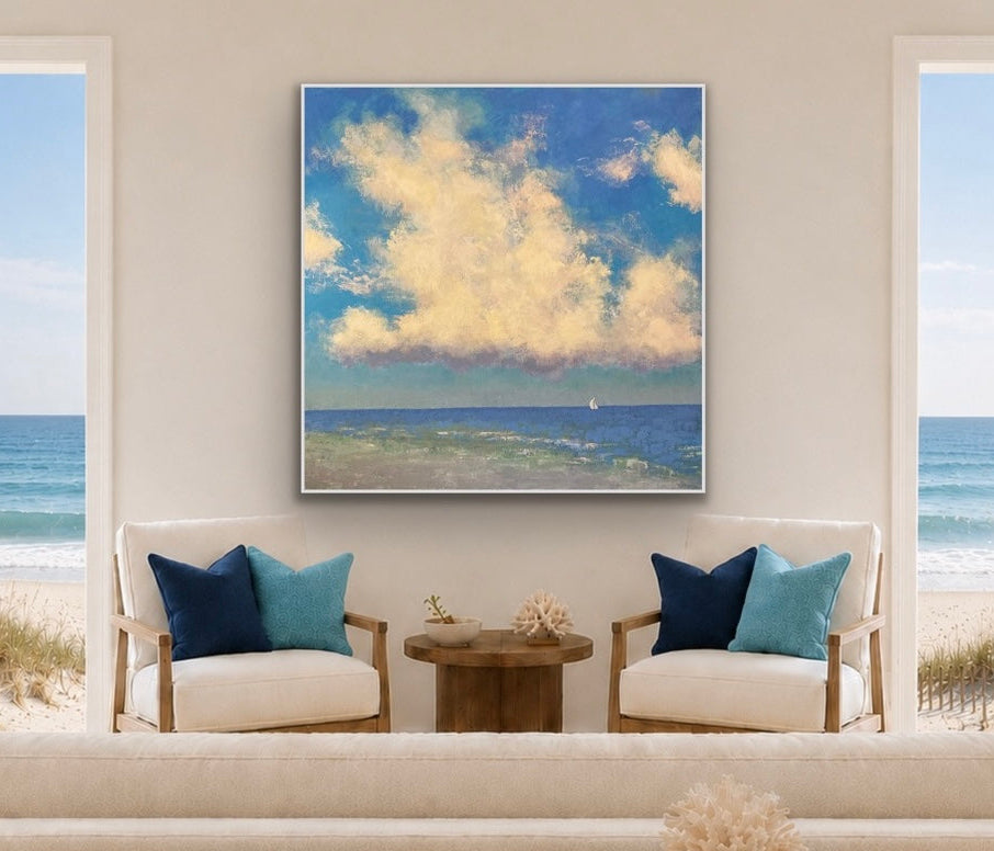 A square framed wall art print depicting a blue ocean and golden clouds, displayed in a bright coastal living room setting above two armchairs with blue and teal pillows.