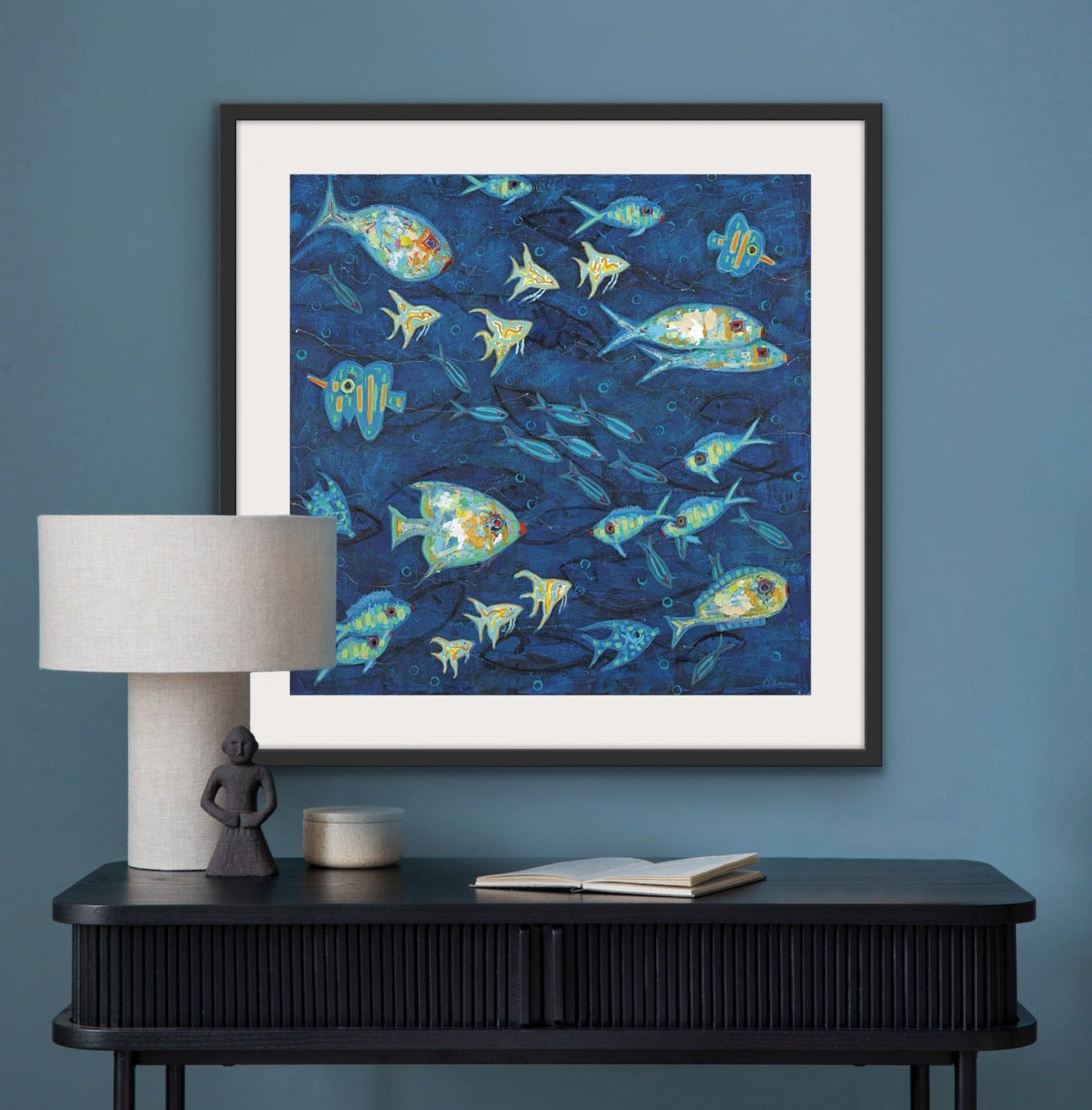 Luxury coastal fish print matted and framed for modern interiors on a blue wall above a black console table with a lamp and stool.