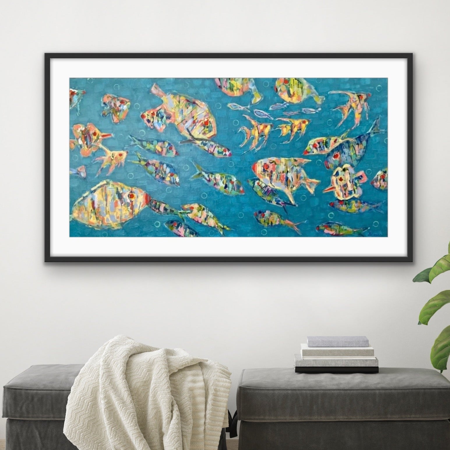 Framed colorful underwater abstracct fish art on a wall above a gray ottoman with a plant to the right.