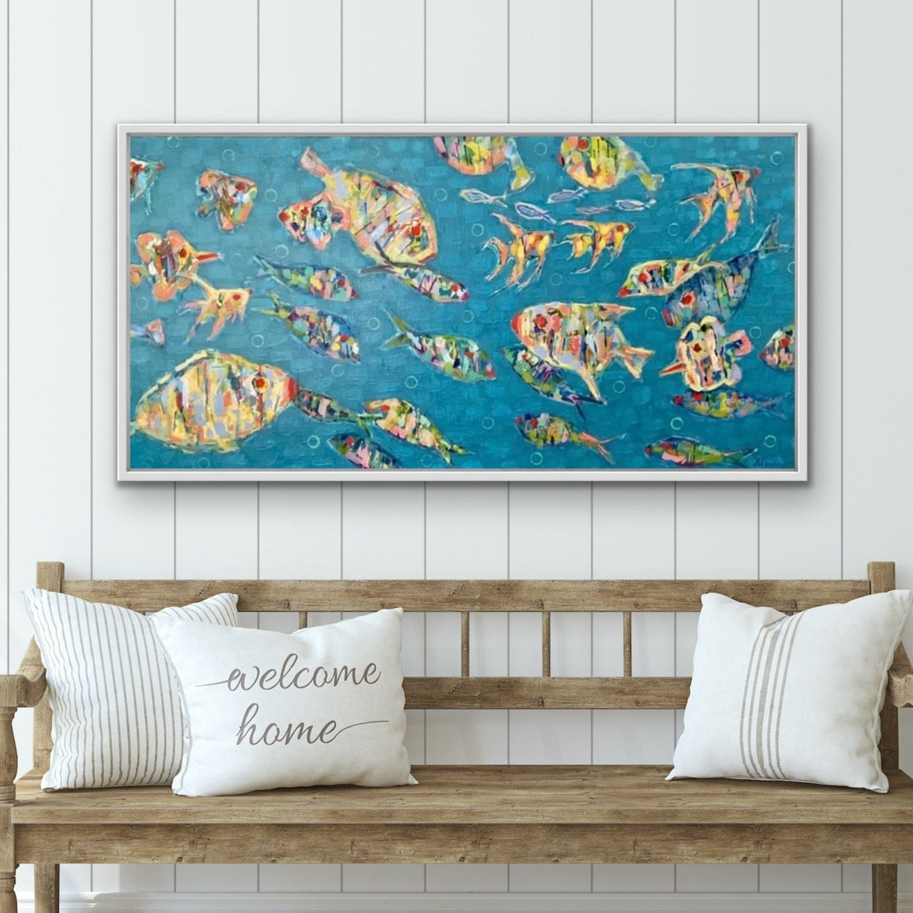 Wooden bench with 'welcome home' pillow, colorful fish painting on wall, and plant in a room.