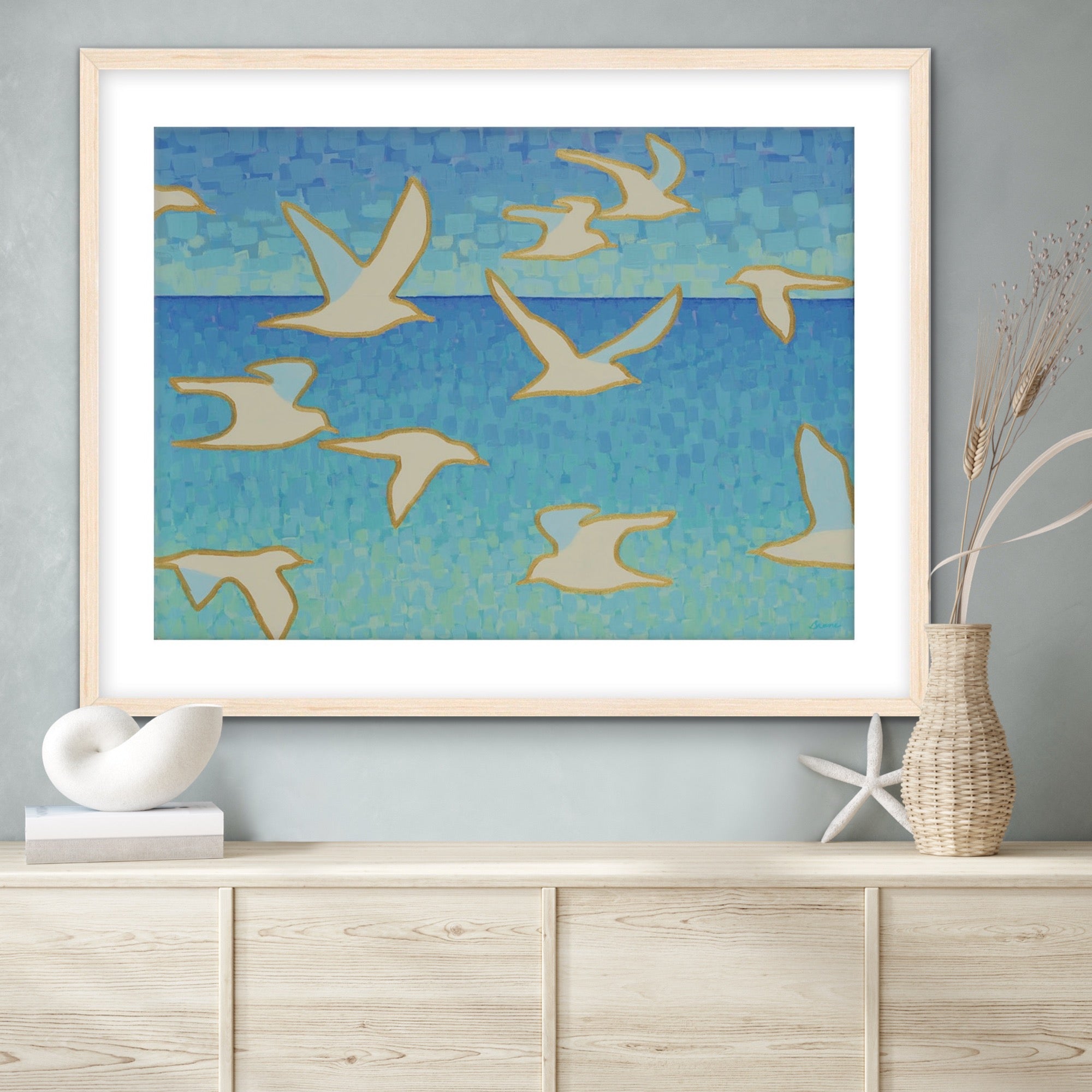 Framed ocean-inspired artwork of birds flying over water on a wall above a wooden cabinet.