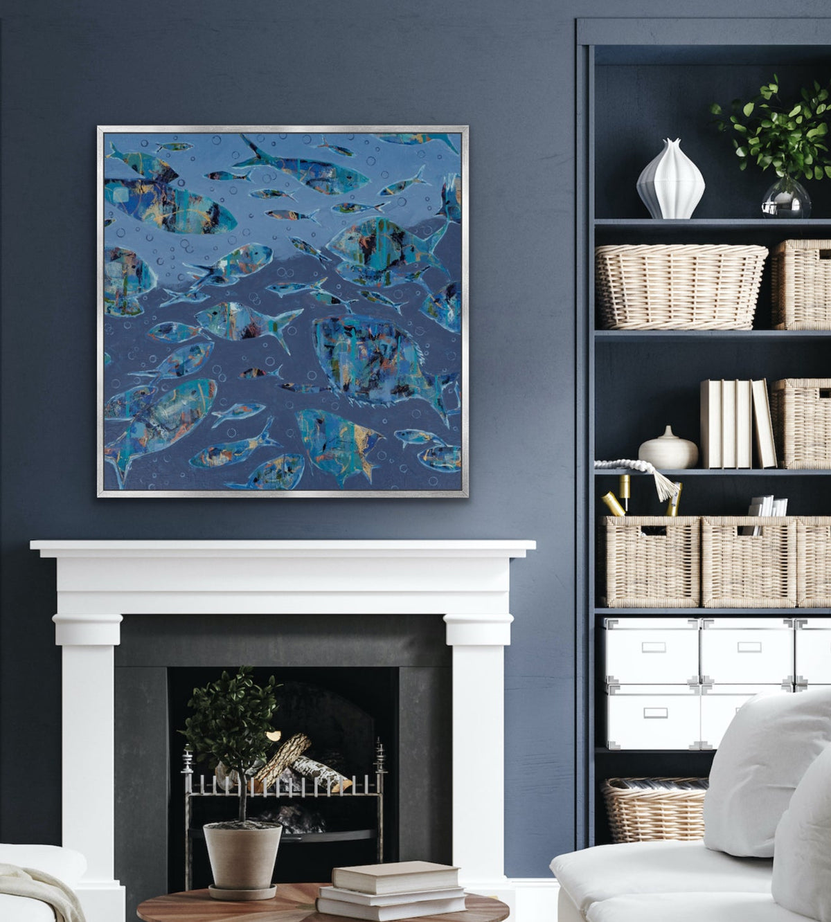 A framed abstract painting with swirling blue, teal, and navy fish patterns hangs on a dark blue wall above a white fireplace mantel in a styled living room with a sofa, coffee table, and shelving.