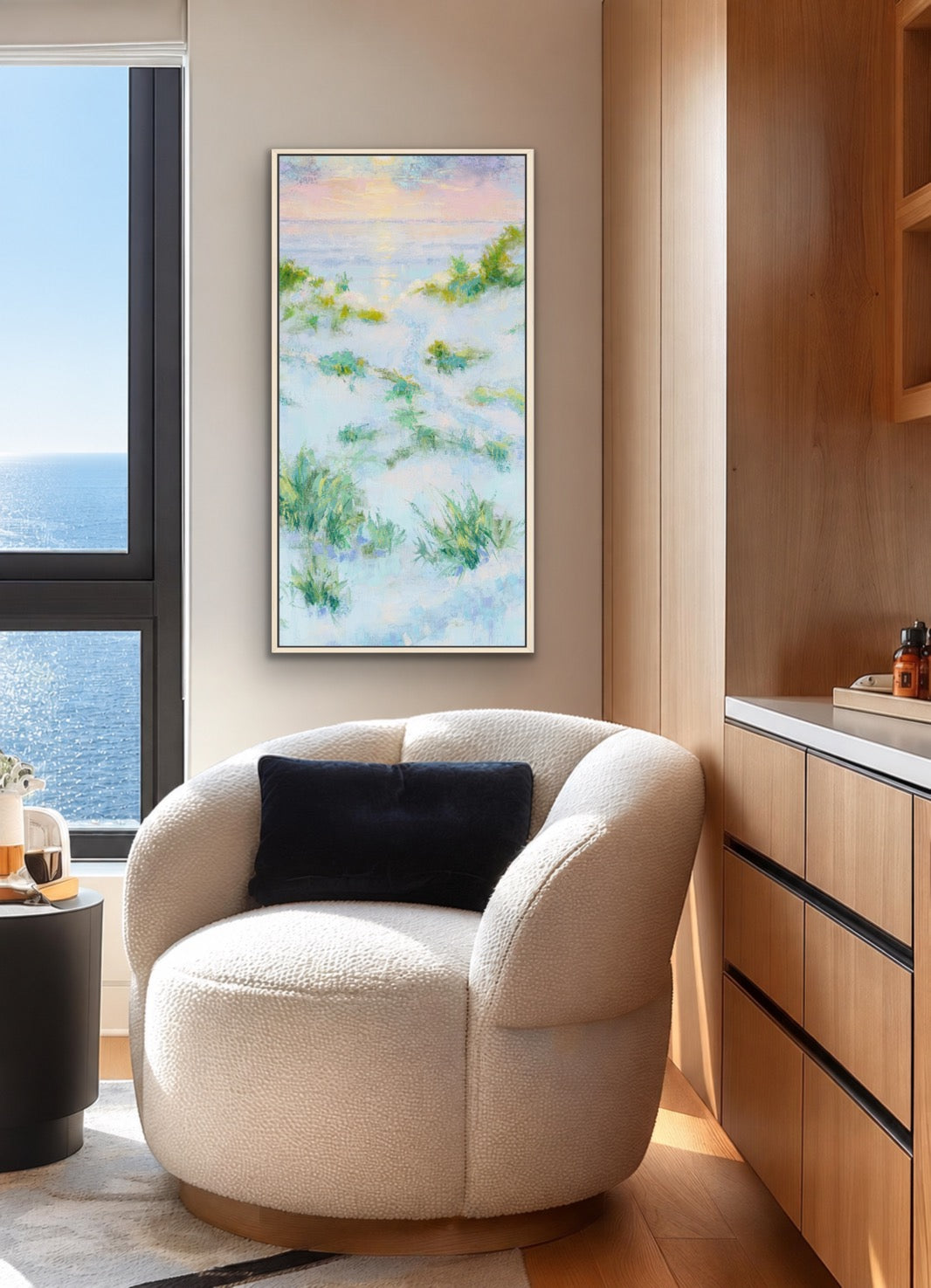 A framed abstract landscape painting with soft blue, green, and yellow tones hangs on a beige wall in a modern room with a cream boucle armchair, a black pillow, and a wooden cabinet.