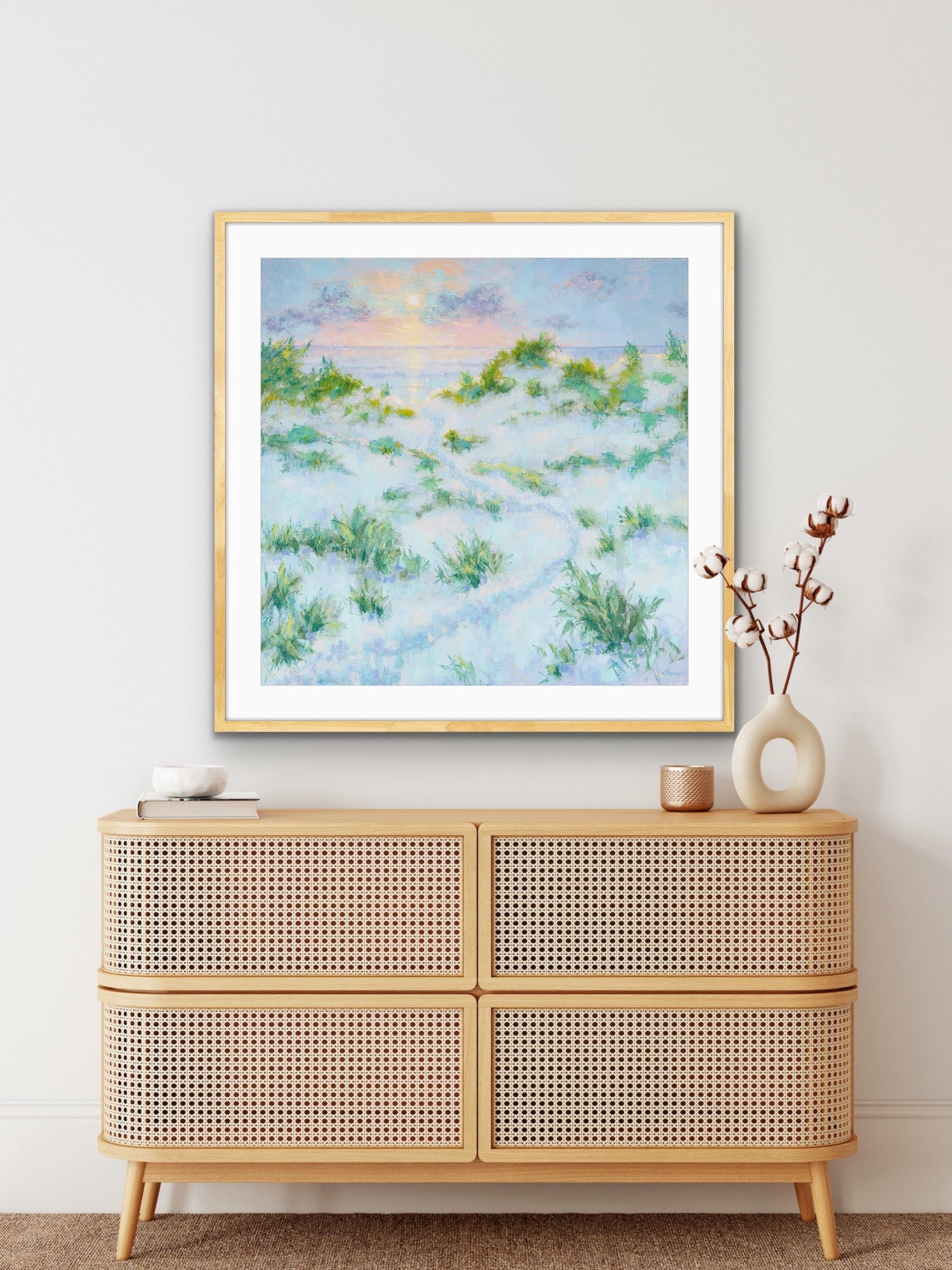 Framed artwork of a sunrise dune beach scene above a rattan cabinet with decorative items.