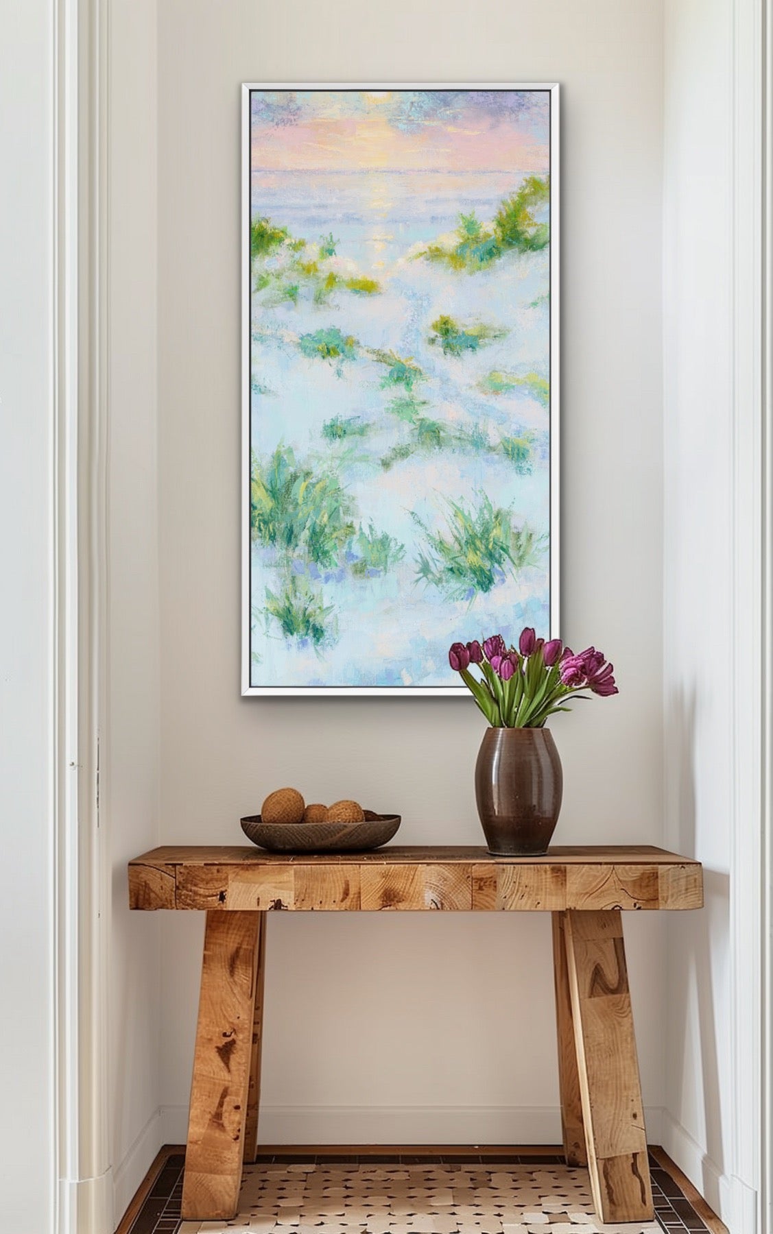 A vertical framed art print featuring an impressionistic beach or marsh landscape in soft blue, green, and pastel tones, displayed on a beige wall above a rustic wooden console table with a vase of purple tulips.
