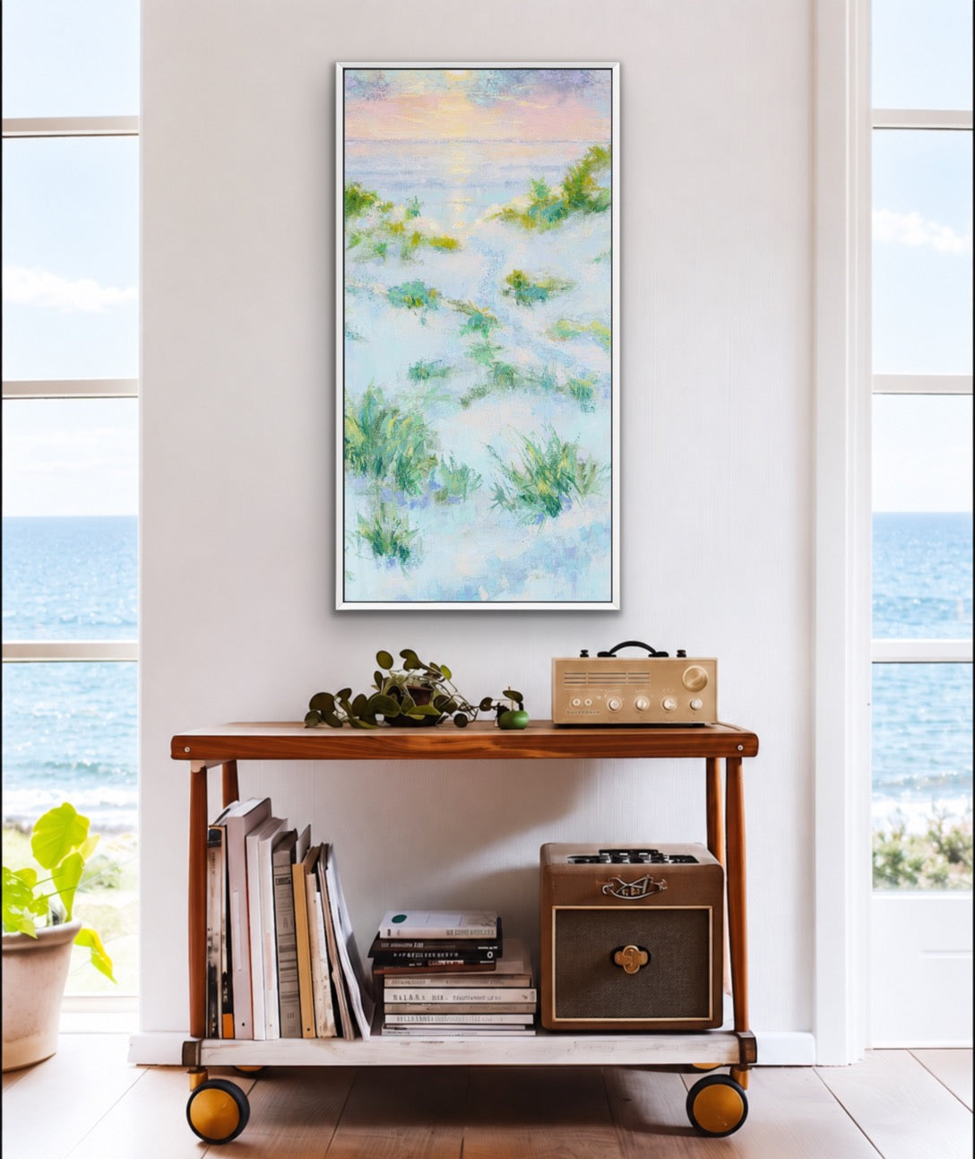 A vertical framed painting of a beach scene with green dune grasses and a pastel sky, displayed on a white wall above a wooden rolling cart in a bright room with ocean views.