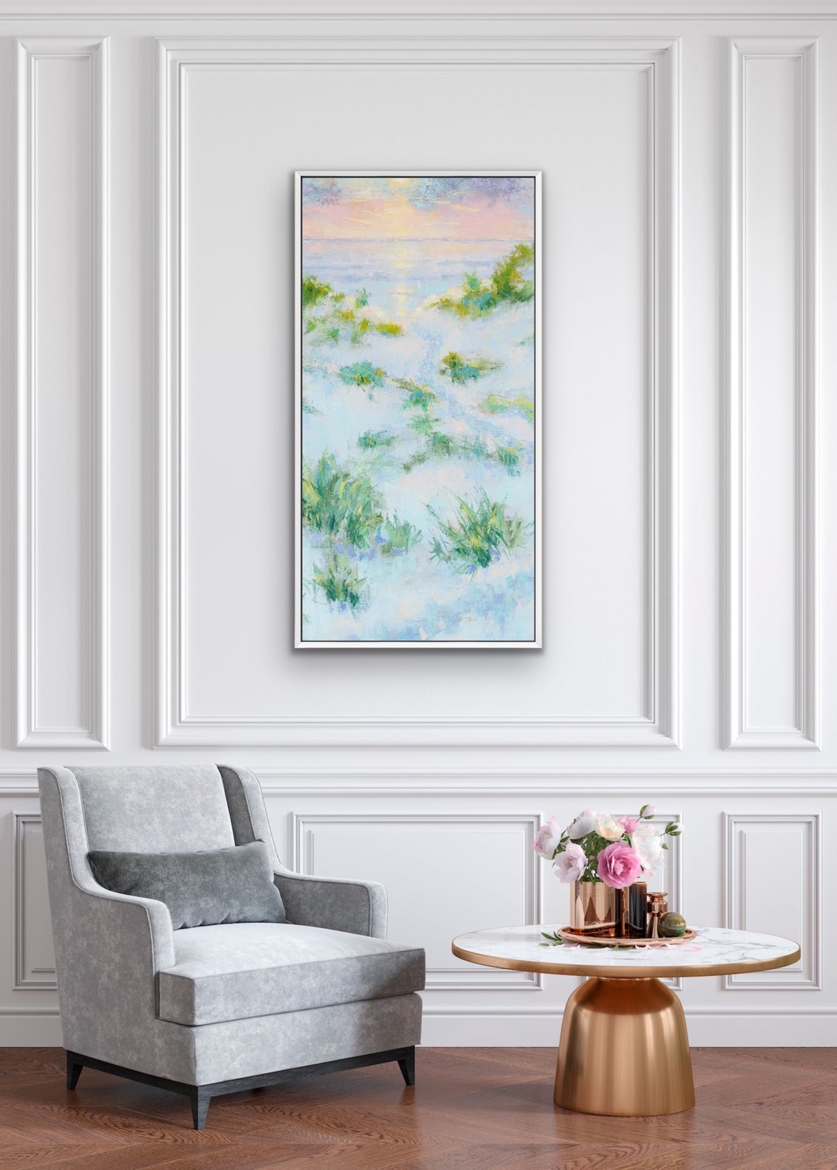A vertical framed painting with an impressionistic beach or grassy landscape in soft blue, green, and yellow tones, displayed on a white paneled wall above a gray armchair and a round gold side table with flowers.