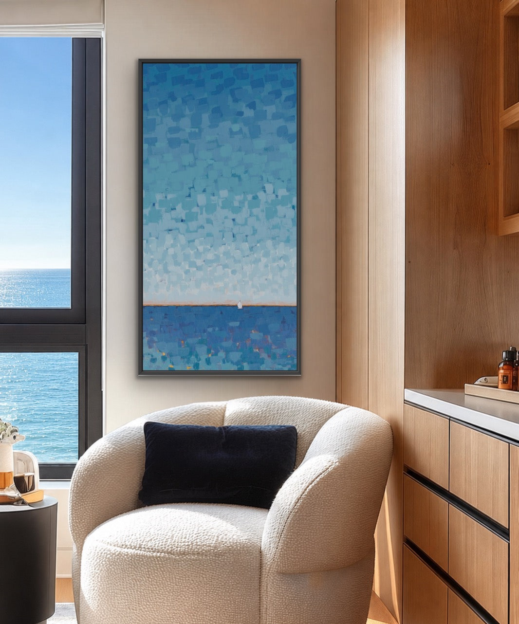 A vertical framed painting of a blue ocean scene hangs on a beige wall in a modern room with a cream boucle armchair, a dark throw pillow, and a wooden cabinet.