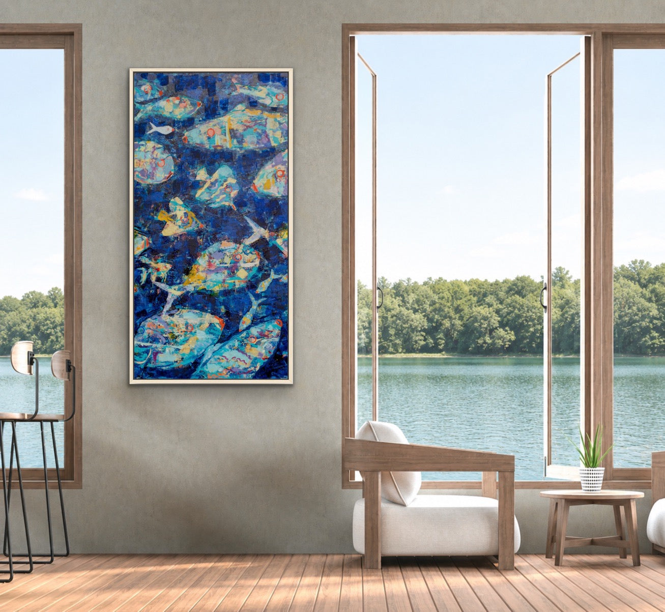 A vertical framed abstract painting featuring blue, teal, and yellow fish motifs, displayed on a gray wall in a modern room with large windows overlooking a lake.